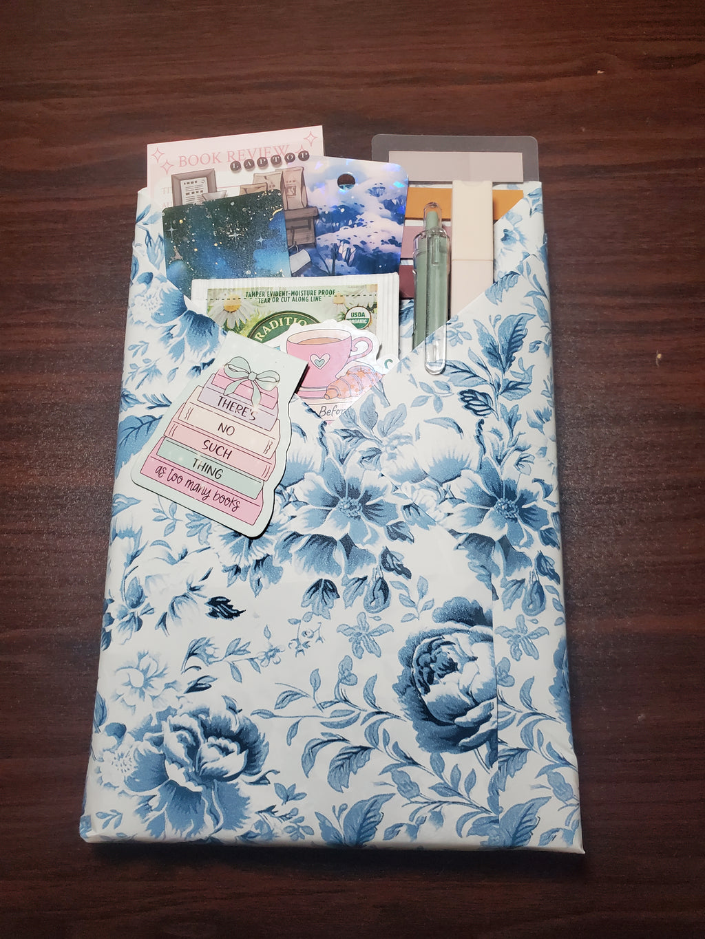 Close-up of a Blind Date With a Book gift wrapped in blue and white floral paper, showing bookmarks, tea, and a book-lover sticker that reads 'There's no such thing as too many books'
